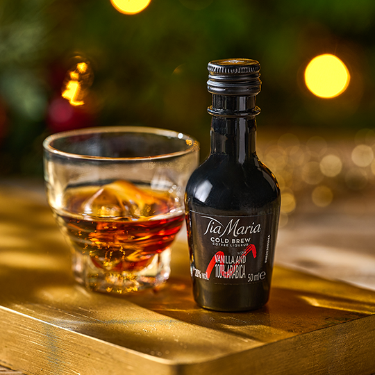 A miniature bottle of Tia Mari beside a small glass with ice in it against a festive background