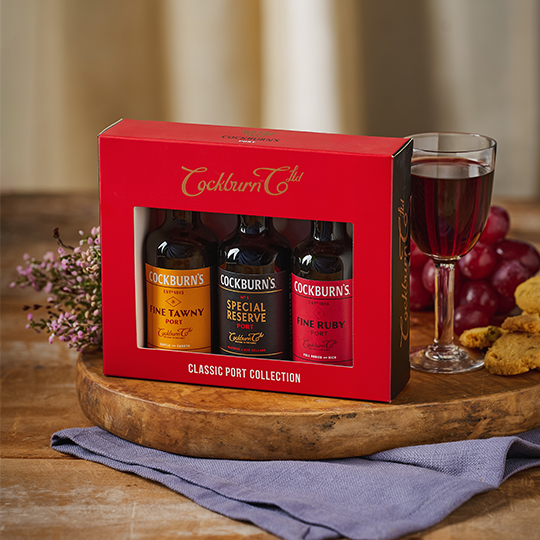 £ small bottles of port in a red gift box, with a glass of port and some grapes to the side
