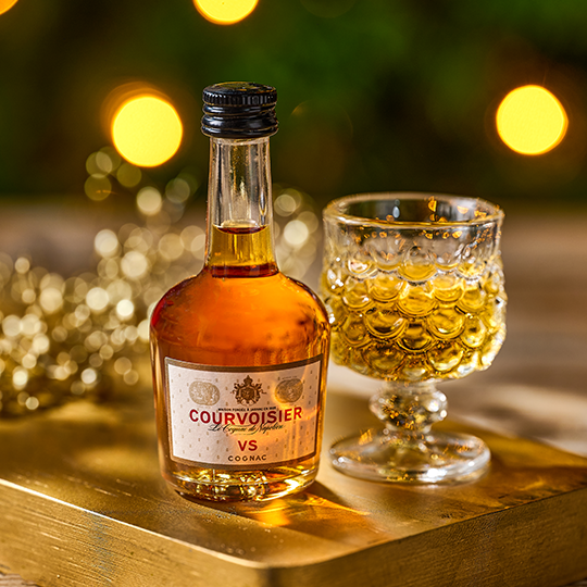 A miniature bottle of Courvoisier beside a small glass against a festive background