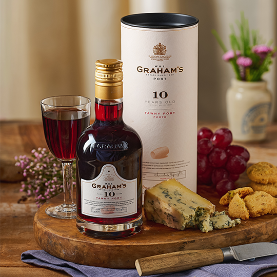 A bottle of port on a board with a wedge of blue cheese, grapes and a savoury biscuit