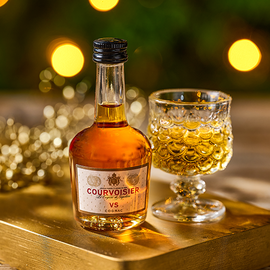 A miniature bottle of Courvoisier beside a small glass against a festive background